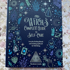 May include: A hardcover book titled "The Witch's Complete Guide to Self-Care" with a dark blue cover. The cover features illustrations of mystical symbols in teal, purple, and white. The book's subtitle reads "Everyday Healing Rituals and Soothing Spellcraft for Well-Being."