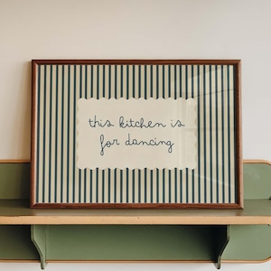 May include: A framed print with the text "this kitchen is for dancing" sits on a green and tan shelf. The print has a brown frame and a background of vertical stripes. The shelf is mounted on a white wall.
