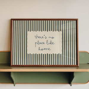 Może przedstawiać: Oprawiony wydruk z napisem "there's no place like home" stoi na zielono-beżowej półce. Wydruk ma brązową ramę i pasiasty wzór w tle. Półka jest zamontowana na białej ścianie.