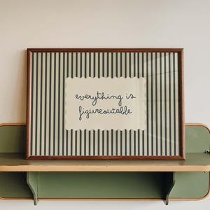 May include: A rectangular framed print featuring dark blue vertical stripes on a cream background with a central scalloped white label containing the black cursive text everything is figureoutable, resting on a sage green wall-mounted shelf.