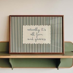 May include: A framed print with the text "actually it's all fun and games" sits on a green and tan shelf. The print has a brown frame and a striped background. The shelf is mounted on a white wall.