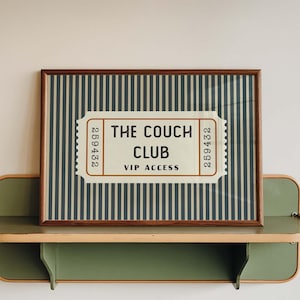May include: A rectangular framed print featuring a ticket graphic with vertical blue and white stripes. The ticket text reads THE COUCH CLUB VIP ACCESS with the number 259432 on both sides. The print sits on a green and light wood wall shelf.