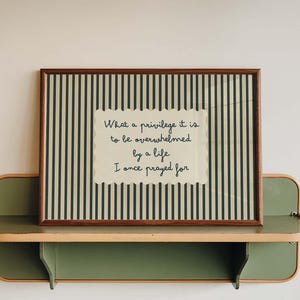 May include: A rectangular framed print with a dark green and cream vertical striped background. Centered is a cream-colored scalloped paper cutout featuring the black text: What a privilege it is to be overwhelmed by a life I once prayed for.