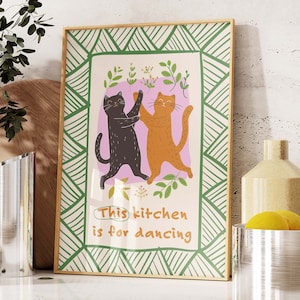 Puede incluir: Una impresión enmarcada con un borde geométrico verde y blanco. La impresión presenta dos gatos bailando con el texto "This kitchen is for dancing".