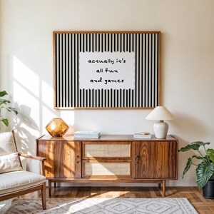 May include: A wooden framed art print with black and white vertical stripes and the text "actually it's all fun and games". The print hangs above a mid-century modern wooden cabinet with a lamp and decorative objects.