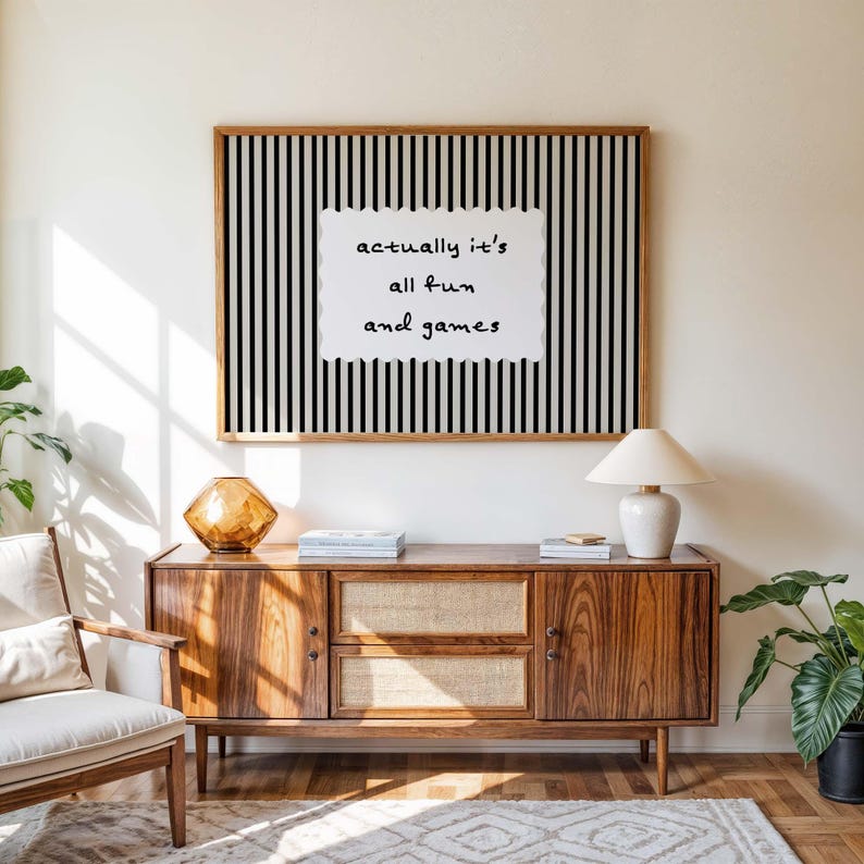 May include: A wooden framed art print with black and white vertical stripes and the text "actually it's all fun and games". The print hangs above a mid-century modern wooden cabinet with a lamp and decorative objects.