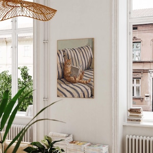 Op de afbeelding: Een ingelijste prent toont een oranje tijgerkat die een boek leest op een gestreepte bank. Het kunstwerk gebruikt een neutraal kleurenpalet, met de kat als middelpunt. De bank heeft blauw-witte strepen. De prent wordt aan een witte muur getoond.
