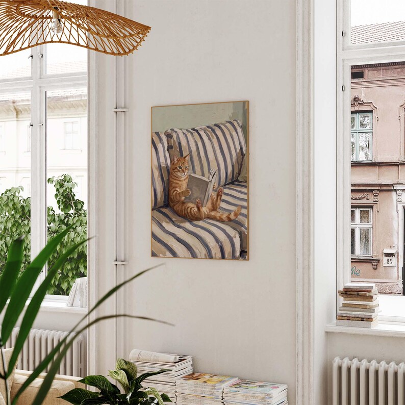 Op de afbeelding: Een ingelijste prent toont een oranje tijgerkat die een boek leest op een gestreepte bank. Het kunstwerk gebruikt een neutraal kleurenpalet, met de kat als middelpunt. De bank heeft blauw-witte strepen. De prent wordt aan een witte muur getoond.