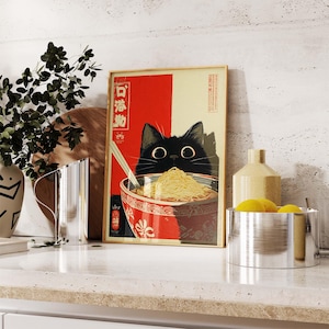 May include: A framed art print featuring a black cat peering over a bowl of ramen noodles. The artwork has a red and cream background with Japanese text. The cat has large, round eyes and is holding chopsticks.