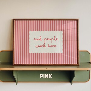 May include: A rectangular framed print featuring a pink and white vertical striped background with a central scalloped white rectangle containing the red text "cool people work here". The frame is dark brown and rests on a green metal shelf.