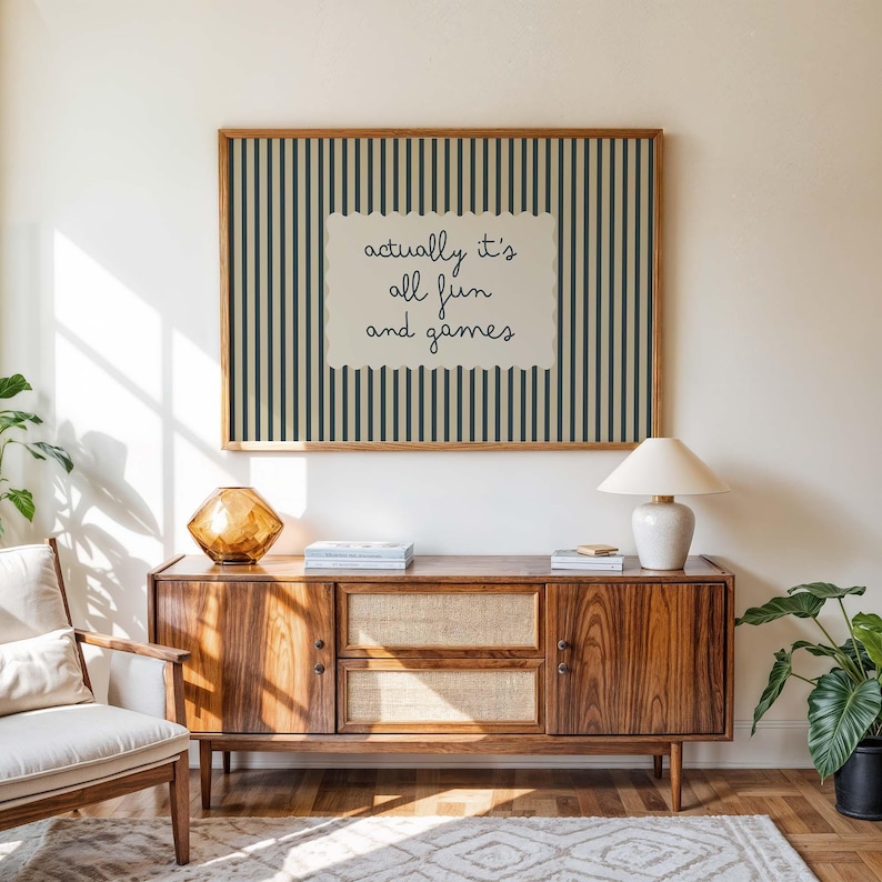 May include: A framed wall art piece with a striped background in dark green and cream colors. The art piece has the text "actually it's all fun and games" in a handwritten style. A wooden cabinet with a lamp and decorative items is below the art.