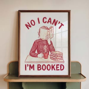 May include: Framed art print with a red and beige illustration. The image shows a person reading a book, a mug, and a stack of books. The text "NO I CAN'T I'M BOOKED" is written in red.