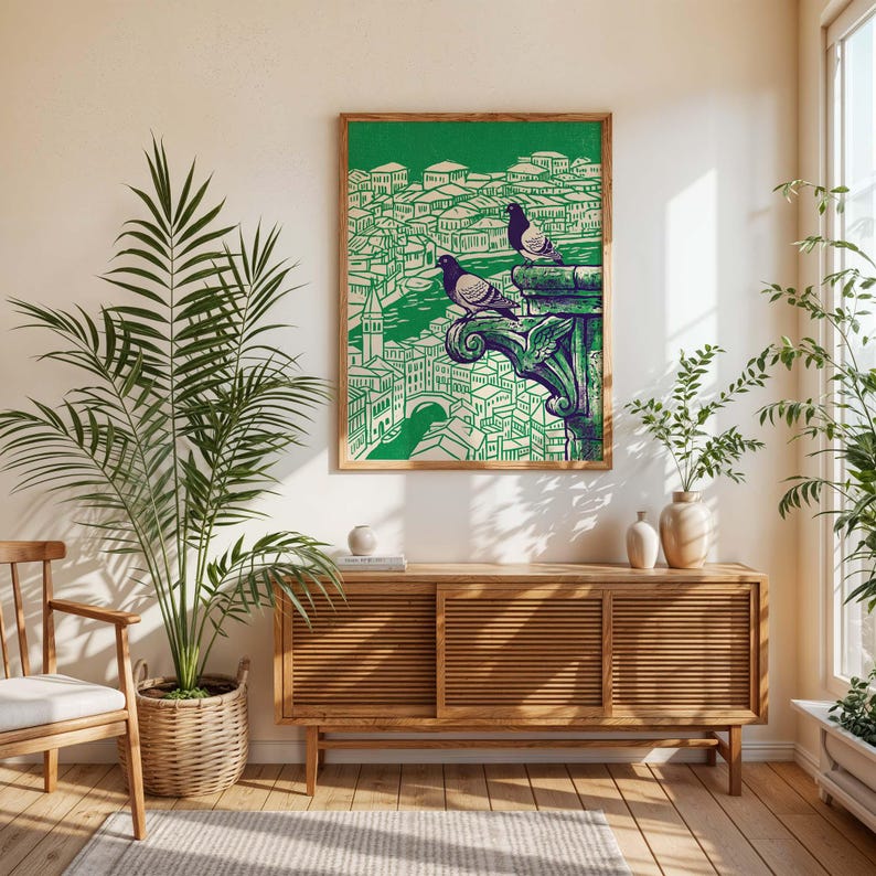 May include: A framed art print featuring a green and cream illustration of a cityscape with two pigeons perched on a decorative architectural element. The artwork is in a wooden frame and hangs above a wooden cabinet.