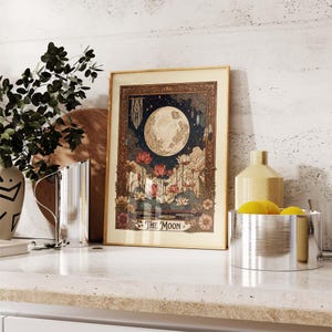 May include: A framed art print with a celestial design. It features a large moon, lotus flowers, and the text "THE MOON." The artwork has a vintage aesthetic with a gold frame and a dark blue night sky background.