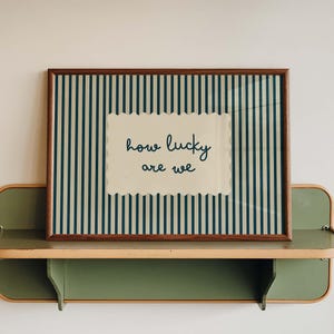 May include: A rectangular framed print featuring a dark blue and cream vertical striped background. In the centre, a cream-coloured scalloped rectangle displays the dark blue handwritten text that reads, how lucky are we, in a simple script font.