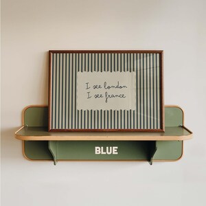 May include: A rectangular framed print with dark blue vertical stripes on a light background, featuring the text I see london I see france in a scalloped white center panel, resting on a sage green wall shelf with the word BLUE in white.