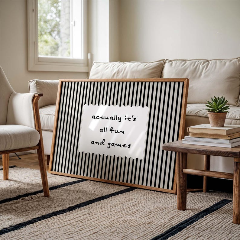 May include: A framed artwork with a black and white striped background and the text "actually it's all fun and games" on a white, torn-edged paper. The frame is made of light brown wood.