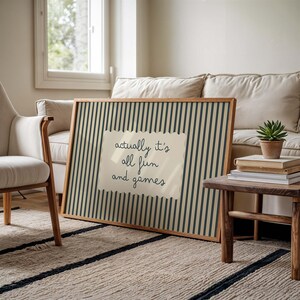 May include: A framed art print with a beige background and vertical navy blue stripes. A cream-colored paper with the text "actually it's all fun and games" is centered on the print. The frame is made of wood.
