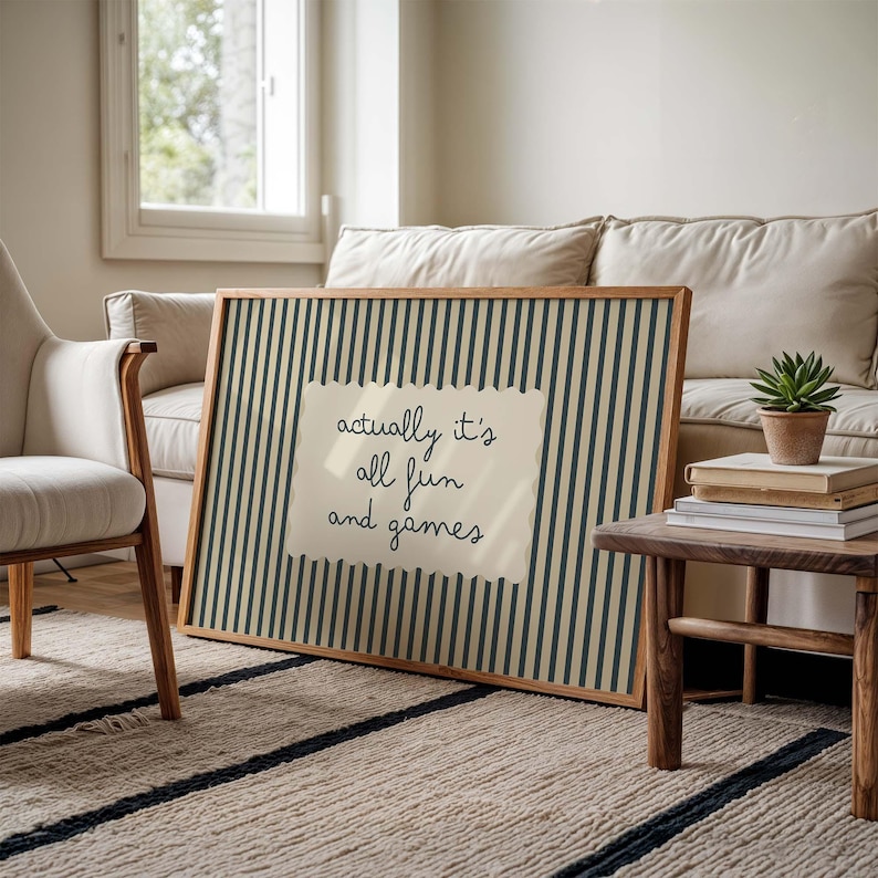 May include: A framed art print with a beige background and vertical navy blue stripes. A cream-colored paper with the text "actually it's all fun and games" is centered on the print. The frame is made of wood.