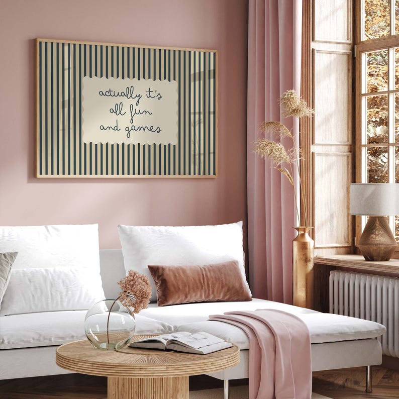 May include: A framed wall art piece with a striped background and the text "actually it's all fun and games" in a living room setting. The room features a white sofa, pink curtains, and a wooden coffee table.