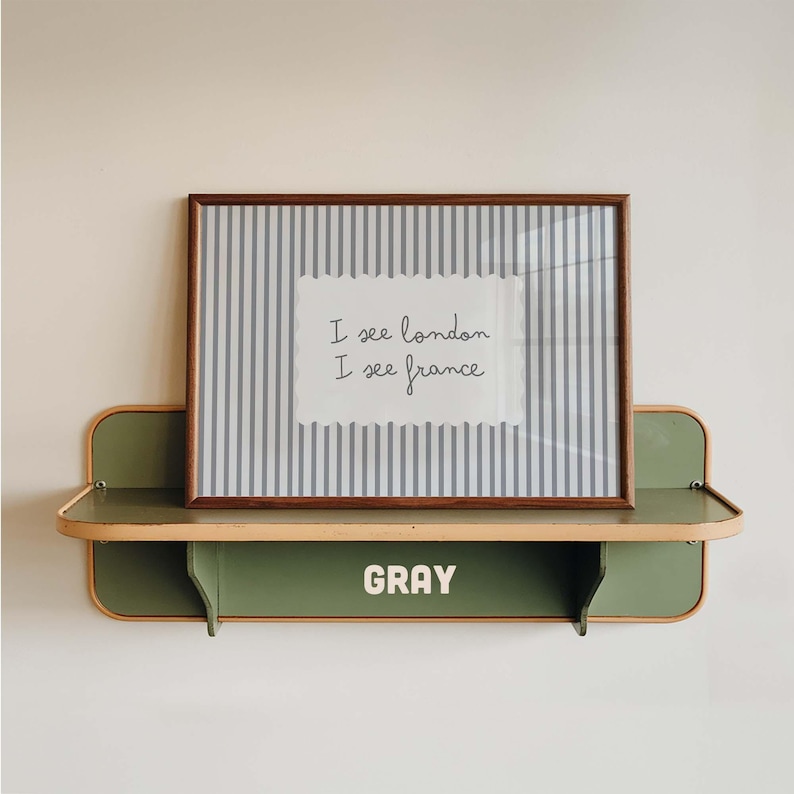 May include: A rectangular framed print with vertical gray and white stripes and a central white scalloped label featuring the black text I see london I see france. The print rests on a sage green metal wall shelf with the word GRAY in white.
