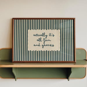 May include: A rectangular wall art print in a dark wood frame featuring a vertical dark blue and cream striped background. Centered is a cream-colored scalloped label with the dark blue text: actually it's all fun and games.
