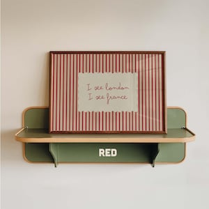 May include: A rectangular framed print with red and white vertical stripes and a central scalloped white panel featuring the red text I see london I see france. The print rests on a sage green metal wall shelf with the word RED printed in white.