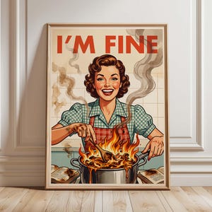 Op de afbeelding: Een ingelijste vintage print met een vrouw in een geruit overhemd en een rode schort, die in een pot met eten op het vuur roert. De tekst "I'M FINE" staat boven haar hoofd. De print heeft een retro esthetiek met een lichtbeige achtergrond.