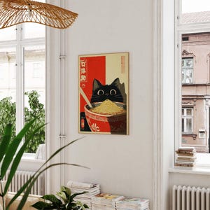 May include: A framed art print featuring a black cat peering over a bowl of noodles. The cat's eyes are large and round. The bowl is red with a white floral design. The background is red and beige with Japanese text.