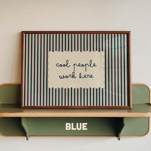 May include: A rectangular wall art print in a dark wood frame featuring a vertical dark blue and cream striped pattern. Centered is a cream-colored scalloped rectangle with the dark blue text "cool people work here" in a cursive font.