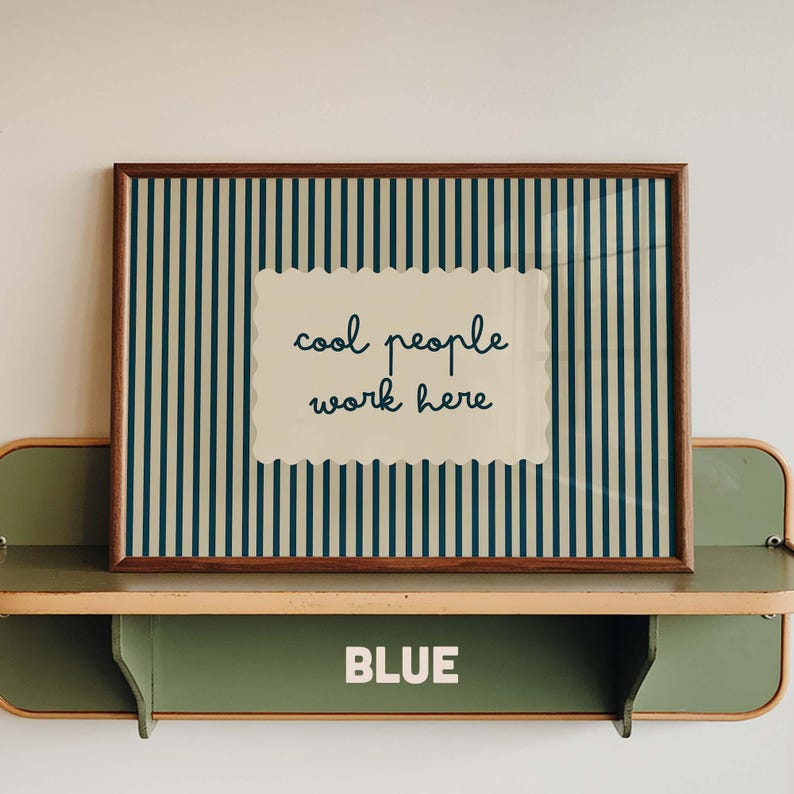May include: A rectangular wall art print in a dark wood frame featuring a vertical dark blue and cream striped pattern. Centered is a cream-colored scalloped rectangle with the dark blue text "cool people work here" in a cursive font.