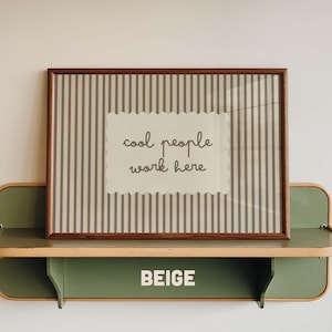 May include: A rectangular wall art print in a dark wood frame featuring a beige background with vertical gray stripes and a central scalloped label containing the dark gray text "cool people work here" displayed on a green shelf.