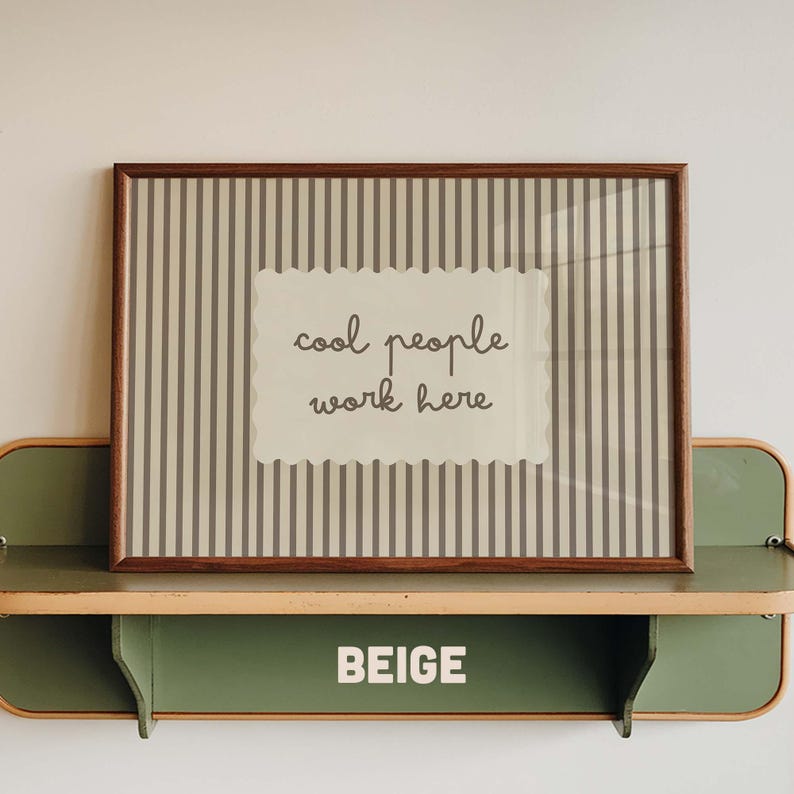 May include: A rectangular wall art print in a dark wood frame featuring a beige background with vertical gray stripes and a central scalloped label containing the dark gray text "cool people work here" displayed on a green shelf.
