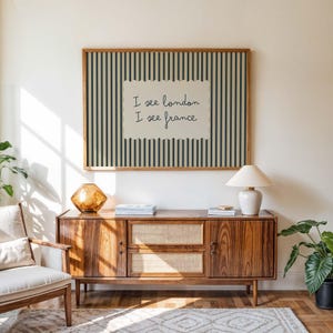 May include: A rectangular wall art piece featuring a dark blue and beige vertical striped background with a central beige scalloped label containing the black handwritten text I see london I see france in a simple wooden frame.