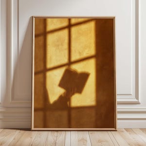 May include: A rectangular wall art print in a light wood frame featuring a warm, golden-yellow scene. The image displays the shadow of a person holding an open book, cast against a textured wall by light streaming through a window pane grid.