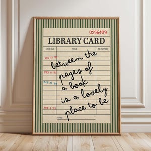 May include: A framed wall art print featuring a vintage library card design with a cream background and dark green vertical stripes. The text reads LIBRARY CARD, 0256489, and includes the handwritten quote between the pages of a book is a lovely place to be.