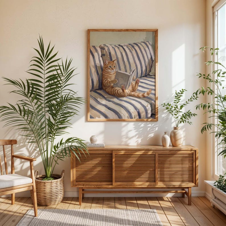 Op de afbeelding: Een ingelijste prent van een oranje kat die een boek leest, zittend op een gestreept blauw en wit kussen. Het kunstwerk hangt boven een houten kast, met planten en een gevlochten mand op de voorgrond.