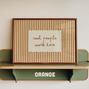 May include: A rectangular framed print featuring vertical brown and cream stripes with a central scalloped-edge cream rectangle containing the brown text "cool people work here". The print rests on a green metal shelf with the word "ORANGE" printed.