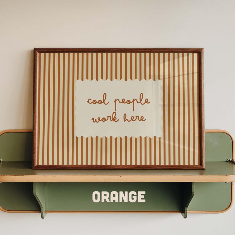 May include: A rectangular framed print featuring vertical brown and cream stripes with a central scalloped-edge cream rectangle containing the brown text "cool people work here". The print rests on a green metal shelf with the word "ORANGE" printed.
