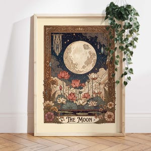May include: A framed art print depicting a full moon, lotus flowers, and a night sky. The illustration has a vintage style with a decorative border and the text "The Moon". The colour scheme includes blues, pinks, and browns. The frame is light-colored.
