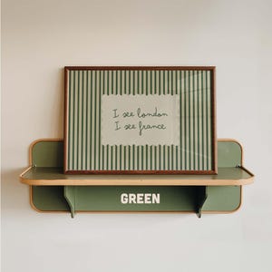 May include: A rectangular wall shelf in muted green with light wood trim, featuring the word GREEN in white capital letters. Resting on the shelf is a framed print with dark green vertical stripes and a central white label reading I see london I see france.