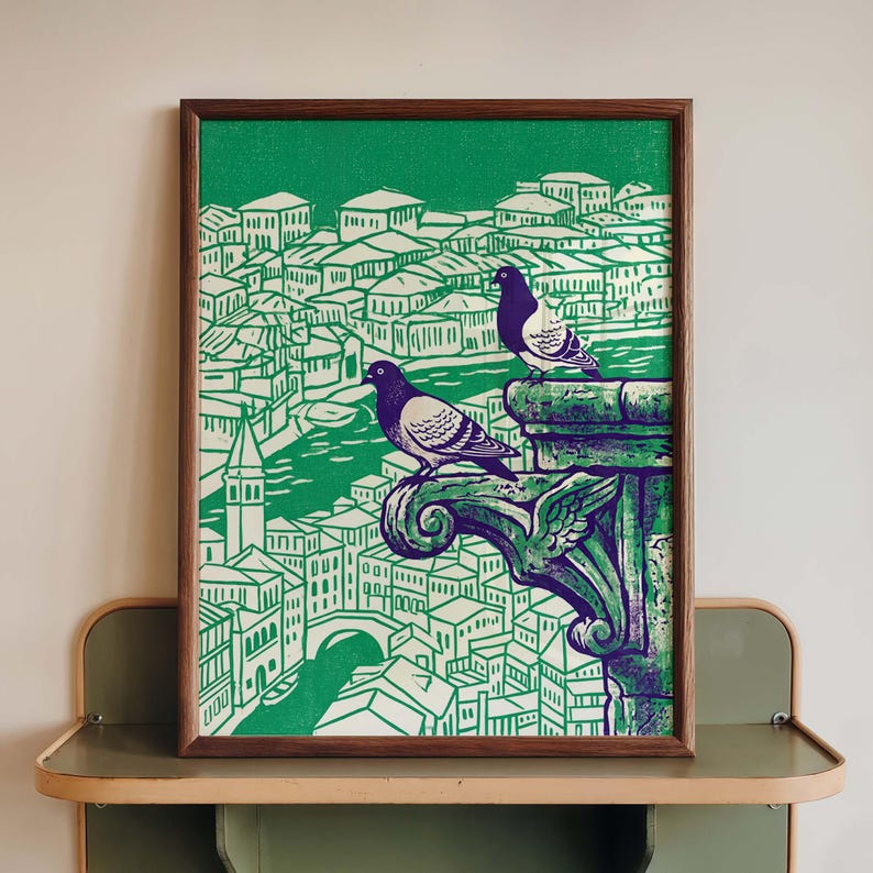 May include: A framed art print featuring a green and white cityscape with two pigeons perched on a decorative stone element. The artwork has a wood frame and is displayed on a shelf. The print has a vintage aesthetic.