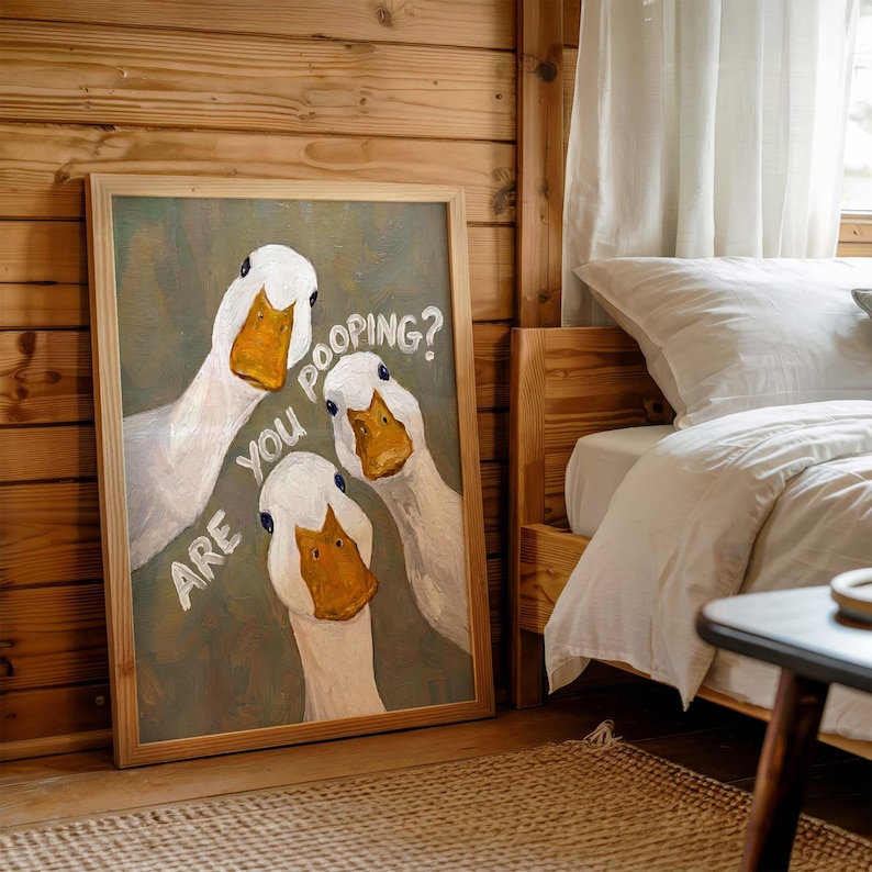 May include: A framed wall art print featuring an oil painting of three white ducks with orange beaks against a muted green background. The text "ARE YOU POOPING?" is written in white, hand-lettered style across the center of the artwork.