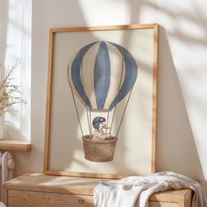 May include: A framed watercolour print featuring a duck in a hot air balloon. The balloon has blue and white stripes, with a brown wicker basket. The duck wears a pilot's helmet and jacket. The frame is light brown wood.