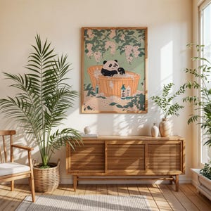 Op de afbeelding: Een vintage-stijl illustratie van een pandabeer die een bad neemt in een houten badkuip, omringd door bloemen en badproducten. De illustratie is ingelijst en hangt aan een muur boven een houten kast.