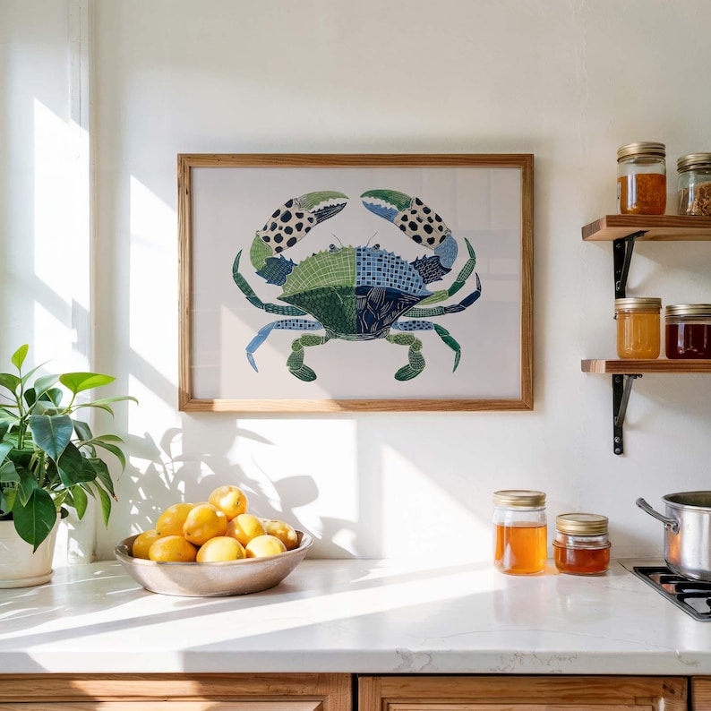 May include: A framed crab art print with a wooden frame. The crab is depicted in shades of green and blue, with patterned details. The artwork hangs on a white wall above a kitchen countertop. A bowl of lemons and jars of preserves are also visible.