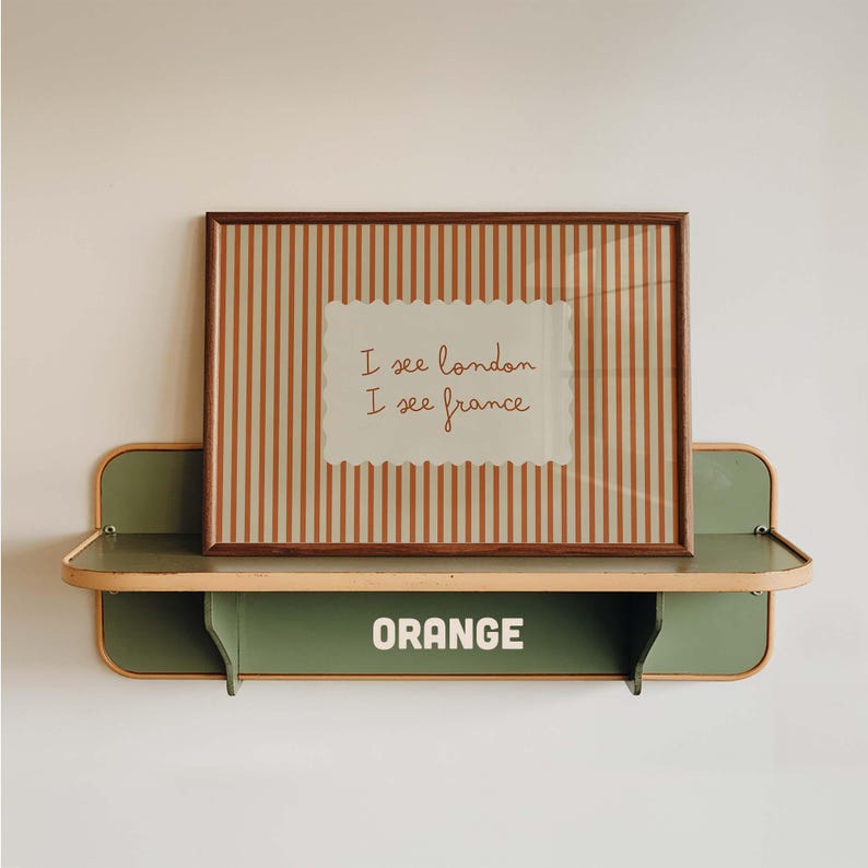 May include: A rectangular framed print with vertical orange and white stripes featuring the text I see london I see france in brown script. The frame rests on a sage green metal wall shelf with rounded edges and the word ORANGE in white text.