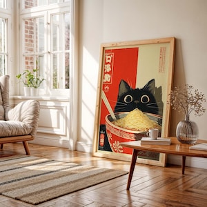 May include: Framed art print featuring a black cat with large eyes peering over a bowl of noodles. The artwork has a red and cream background with Japanese text and is in a natural wood frame. A small coffee cup sits on a wooden table.