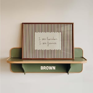 May include: A rectangular framed print with vertical brown and white stripes featuring the text I see london I see france in cursive script, resting on a sage green wall-mounted shelf with the word BROWN printed in white on the front panel.