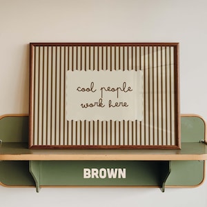 May include: A rectangular framed print featuring a brown and beige vertical striped background with a central scalloped-edge label containing the brown text "cool people work here" displayed in a cursive font style within a dark wood frame.